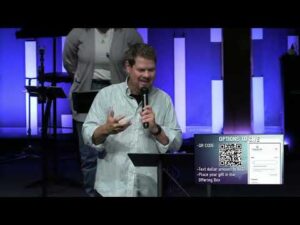 A man speaks at a podium on stage with a microphone. Behind him are lit blue panels and another person. A screen displays options to give, including a QR code and text instructions.