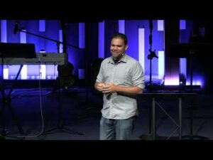 A man stands on a stage in front of musical equipment and blue-lit vertical panels, speaking to an audience.