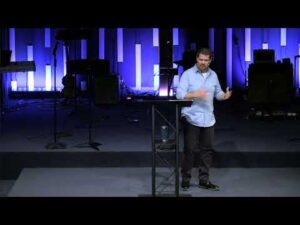 A man stands at a podium on a stage with musical instruments and equipment in the background, illuminated by blue lighting.