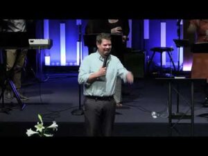 A man stands on stage holding a microphone and speaking, with musical instruments and equipment visible in the background.