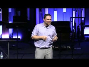 A man in a light purple shirt and gray pants stands on a stage, speaking, with musical instruments and blue-lit panels in the background.