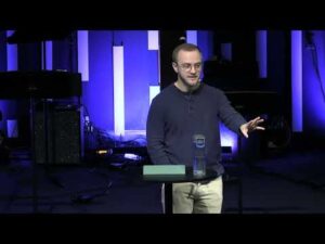A person stands at a round table with a water bottle and book, speaking in front of a stage with vertical light panels and audio equipment.