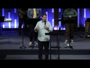 A man stands at a podium speaking to an audience on a stage with three people behind him and blue vertical lights in the background.