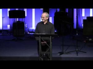 A man in a black shirt stands at a podium on a stage with blue and white vertical lights and musical equipment in the background.