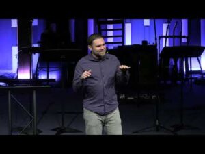 A man stands on stage speaking, gesturing with both hands. Musical instruments and stands are visible in the background, illuminated by blue and purple lights.