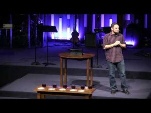 A man stands on a church stage near a wooden table with cups and plates, with musical instruments and stands in the background under purple lighting.