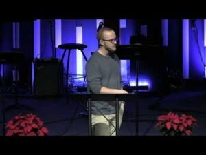 A man stands at a podium speaking in a dimly lit room with vertical blue lights in the background and red poinsettias in the foreground.