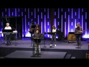 A group of musicians and a speaker stand on a stage with sheet music stands, instruments, and microphones in front of a backdrop lit with vertical blue lights.
