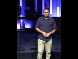 A man stands on a stage in front of musical equipment and vertical light panels, wearing a dark shirt and light pants. The word "LIKE" appears on his shirt.