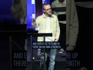 A person in a light jacket stands at a podium speaking, with one hand raised. Text on screen reads: "AND EXPECT US TO CLIMB UP THERE ON HIS OWN STRENGTH.