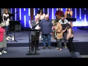 A group of people stands on a stage; a man speaks at a podium while others gather beside him, some holding flowers, with musicians in the background.