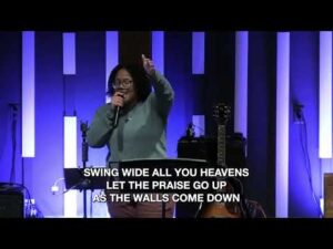 A person stands on stage holding a microphone and raising one hand, with guitars in the background and song lyrics displayed at the bottom.