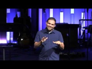 A man in a blue shirt speaks and gestures on a dimly lit stage with musical instruments and vertical light panels in the background.