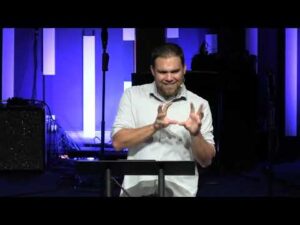 A man stands at a podium speaking, gesturing with his hands, with blue and white vertical lights in the background.