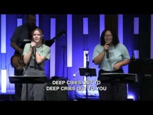 Two people sing on stage with microphones, accompanied by musicians. Projected lyrics read, "DEEP CRIES OUT TO DEEP CRIES OUT TO YOU." Vertical blue and purple lights in the background.