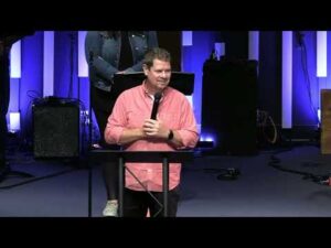 A man in a pink shirt stands at a podium speaking into a microphone in a dimly lit room with blue and purple lighting.