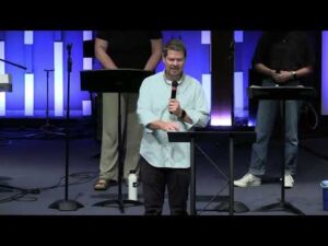 A man speaks into a microphone at a podium on a stage, with three people standing behind him and blue vertical lights in the background.