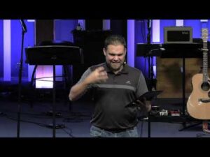 A man stands on stage holding a tablet and gesturing with his hand, with a guitar and musical equipment visible in the background.
