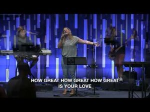 A band performs on stage with blue vertical light panels; a woman sings at the center. Lyrics read: "HOW GREAT HOW GREAT HOW GREAT IS YOUR LOVE.