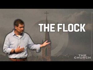 A man gestures while standing in front of an image of a church steeple with the text "THE FLOCK" and "THE CHURCH" displayed on the background.